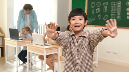 Asian boy looking at camera while waving hands and standing at blackboard. Skilled child greeting...
