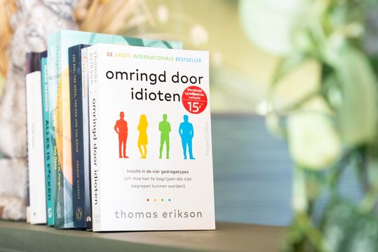 Thomas Eriksons bestseller 'Surrounded by Idiots' (pictured is Dutch version Omringd door Idioten) displayed leaning against a row of books, surrounded by a beautiful bokeh environment.