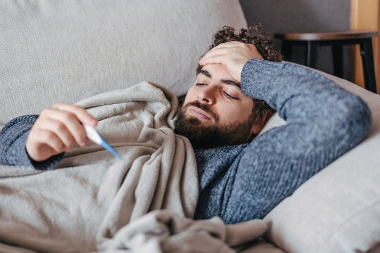 Man with fever checking temperature while lying sick on a couch. cold, flu, virus season