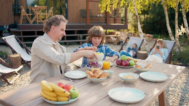 Caring Caucasian father sitting at table together with his son. Passing plate with chips. In background woman sitting together with daughter. Having positive conversating on relaxing day.