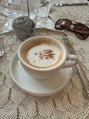Cappuccino topped with cinnamon served on a luxurious beaded tablecloth, accompanied by sunglasses, napkins, and silverware, elegance and sophistication, concept of lifestyle, dining, luxury