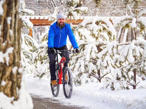 A joyful man rides a bicycle in a winter park among snow-covered trees. Bearded cyclist in a blue jacket.. Active lifestyle