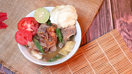 Closeup of a bowl of Beef ribs soup or sop Iga with beef bone, potato, fresh tomato, lime slices, and fried shallot. Served on bamboo mats, burlap, and leaves. Space for text available
