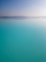A turquoise tailings pond near a cement factory, surrounded by rugged hills. The vivid water contrasts with the barren land, creating a striking yet industrial scene of environmental impact