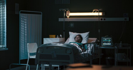Young man sits in the hospital room beside an unconscious girl lying in the bed.