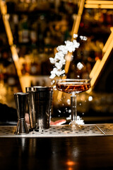 Stemmed glass with a brown cocktail and two parts of a shaker stand on a bar counter