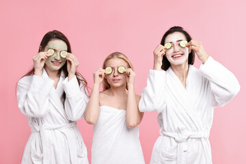Spa day. Happy women with face masks and cucumber slices on pink background