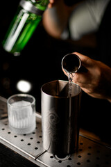 Bartender pours a clear liquid from a jigger into a tall metal glass for mixing a cocktail