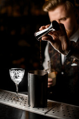 Male bartender pouring the last drops of liquid from a cocktail jigger into a metal glass in a thin stream