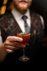 Male bartender holds out a wine glass filled with a brown cocktail