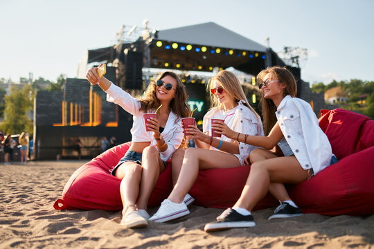 Group of female friends enjoy summer music festival on sandy beach, sitting on red bean bag chairs, sipping cool drinks, stage in background. Happy women in casual attire relax, laugh.