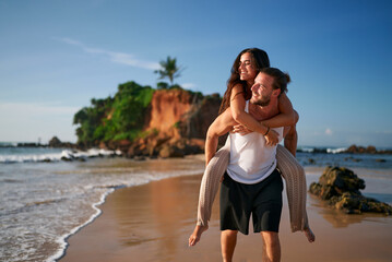 Romantic couple enjoys honeymoon on tropical beach. Man gives piggyback ride to smiling woman at sunset. Newlyweds walk by sea, love moments on exotic island. Happy bride, groom in casual wear.