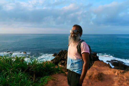 Senior ethnic man hiking near ocean waves. Bearded elderly enjoys adventure travel at rocky coast. Active retiree with backpack explores nature. Outdoors lifestyle in golden years.