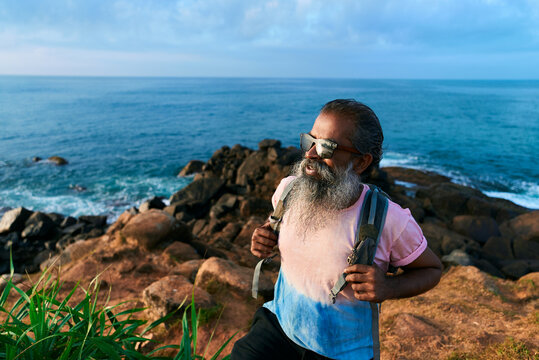 Stylish senior man with beard hikes rocky seaside, wearing sunglasses, backpack. Active adventure. Retirement travel, coastal destination. Healthy, energetic grandpa embracing outdoor experiences.