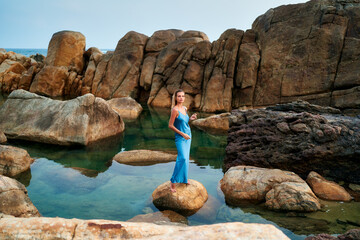 Stunning model in wet blue dress stands on rock at exotic ocean beach. Luxury fashion photoshoot....