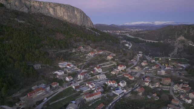 Scenic aerial view of the village of Klis, nestled on a hillside with forested areas, traditional houses, and a highway visible in the background under a serene evening sky.