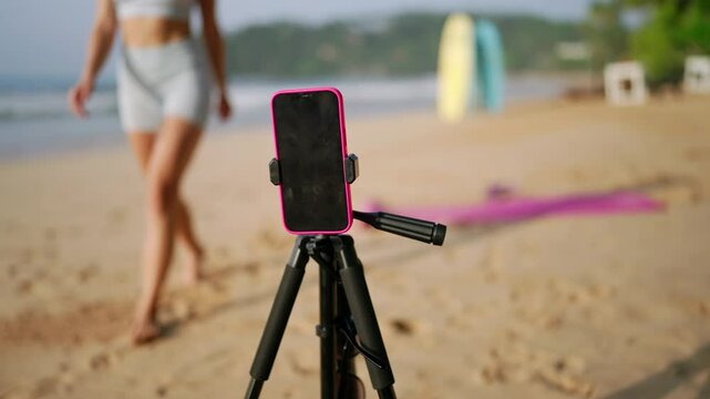 Fitness instructor conducts beach workout session using smartphone on tripod, live streams exercise routine, interacts with virtual attendees, demonstrates moves against tranquil seaside backdrop.