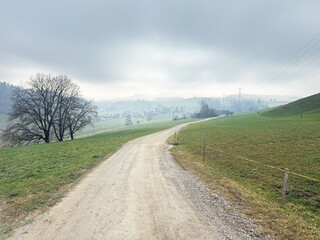A dirt road stretching through a green countryside landscape under a cloudy sky. Power lines are visible on the left, adding contrast to the natural surroundings. The scene captures the calm and