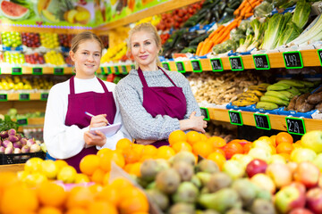 Portrait of confident smiling saleswoman standing with trainee salesgirl among shelves with fresh organic fruits and vegetables in grocery store