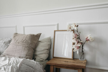 Blank wooden picture frame mockup on wooden side table with glass vase. Blooming magnolia tree branches. Scandinavian interior. Elegant bedroom, beige pillows. White wall background, stucco decor.