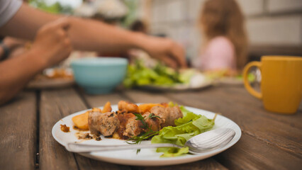 Rustic wooden table displaying hearty leftovers, grilled sausages, roasted potatoes, green salad on white plate, blurred family dining scene behind