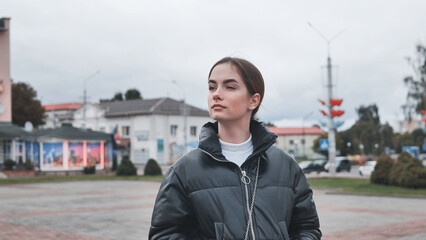 Stylish young woman wearing black puffy jacket and white turtleneck enjoys a cloudy day in a town...