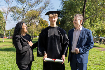 Male graduate dressed in cap and gown celebrating on campus with his multi-ethnic family