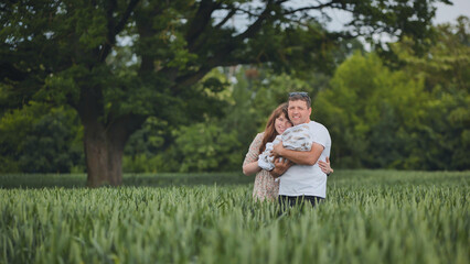 Parents holding their newborn baby in a green wheat field under a big tree, enjoying a peaceful family moment in nature