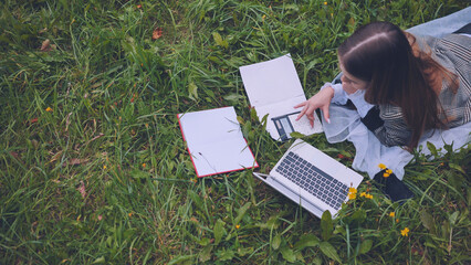 Schoolgirl lying on lush grass, using laptop and calculator while completing homework outdoors,...