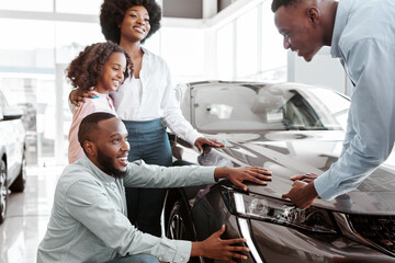 Fototapeta premium Car sales business. Young black family with cute child selecting vehicle, looking at luxury automobile at showroom. Seller talking to Afro clients, showing them new auto at dealership shop