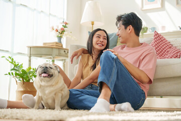 Asian couple sitting on the floor in a living room, smiling warmly while a pug dog sits beside them, with sunlight through large windows
