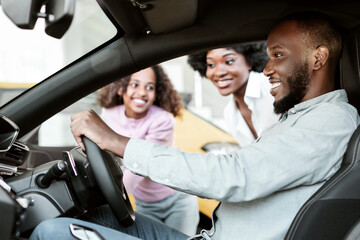 Happy black guy sitting in new car, test driving automobile, choosing auto with his family at dealership, panorama. Afro father with his wife and daughter selecting vehicle at showroom store