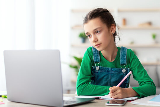 Web-based Distance Education Concept. Portrait of diligent teen sitting at table in living room, using looking at laptop screen writing in her notebook. Schoolgirl watching online course, taking notes