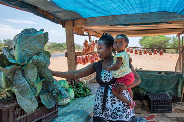 African woman vendor holding baby selling vegetables from a wooden shack stall in the city, on the...