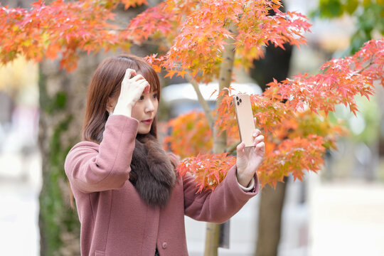 In front of autumn leaves. Japanese women with long hair in their 30s take a selfie with a smartphone. - Powered by Adobe