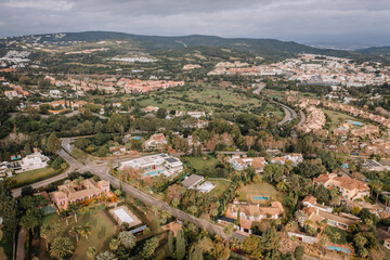Obraz premium Aerial view of a luxury residential area with villas, private pools, gardens, and nearby greenery surrounded by scenic hills and urban landscapes.
