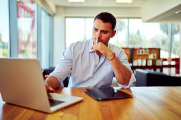 Obraz premium Businessman concentrating and thinking while working on laptop in modern office, with tablet on wooden desk
