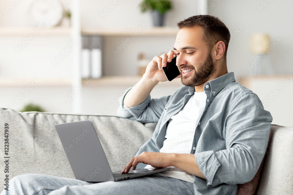Aufkleber attractive young guy talking on smartphone and using laptop computer on couch at home office. handso
