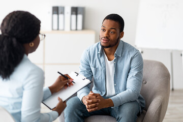 Unhappy young black man having session with professional psychologist at mental health clinic. Psychotherapist taking notes during conversation with depressed male patient