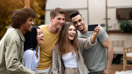 Positive multiracial friends taking selfie near their trailer, smiling and having fun together during autumn camping trip. Cheerful young people making photo of themselves at campsite