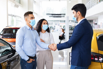 Obraz premium Middle-eastern millennial couple in protective face masks shaking salesman hand, celebrating successful deal, buying new car in brand new showroom while coronavirus pandemic, side view