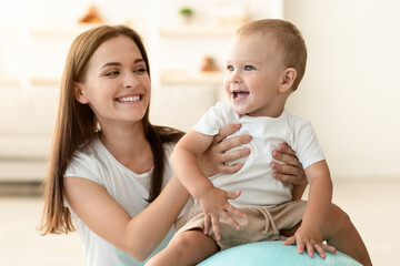 Happy Young Mother And Cute Little Baby Playing With Fitness Ball At Home, Practicing Sport Together, Adorable Infant Boy Sitting On Fitball And Laughing, Enjoying Spending Time With Mom, Closeup
