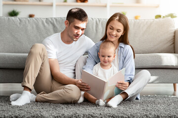 Development Leisure For Kids. Happy Parents Reading Book With Toddler Son At Home, Millennial Family Of Three With Infant Child Relaxing On Floor In Living Room, Enjoying Domestic Leisure, Copy Space