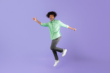 Obraz premium Joyful black guy jumping up in the air and showing funny figures, showing positive emotions over blue background, crop. African american teenager posing at studio, having fun