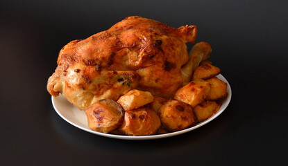 Large round plate with whole baked chicken and new potatoes on black background.