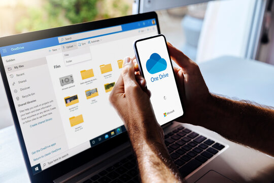 OneDrive on mobile phone and computer. Man holding a mobile phone with Microsoft cloud storage app on the screen. Laptop at background showing One Drive folfders. Rosario, Argentina - January 27, 2025