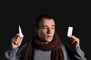 Ill young man with nasal drops and pills on dark background, closeup
