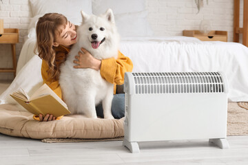 Young woman with cute dog reading book near radiator in bedroom