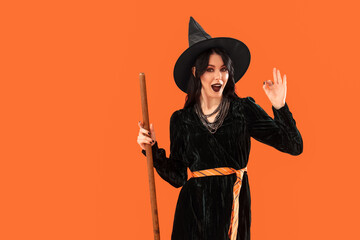 Beautiful young woman dressed as witch for Halloween with broom showing OK gesture on orange...