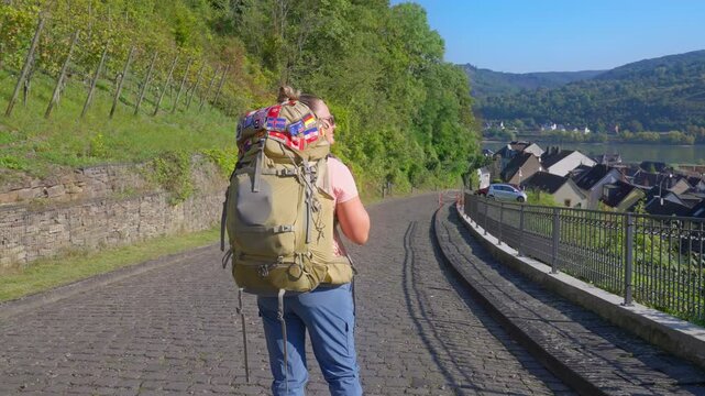 Female trekker with backpack filled with country patches taking in Rhine River Valley view over a small German town and rolling hills in the distance on a sunny summer morning